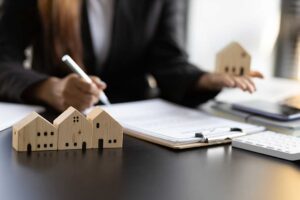 An appraiser reviews a house exterior while holding a clipboard and comparing property details with sales records.